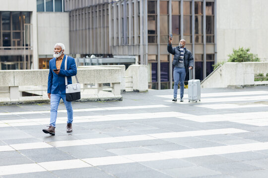 Senior african american men walking on plaza checking phones and pulling suitcase