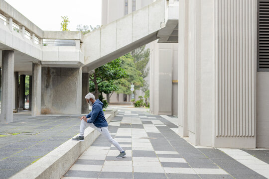 African american senior man stretching on concrete ledge outdoors in hoodie and earbuds