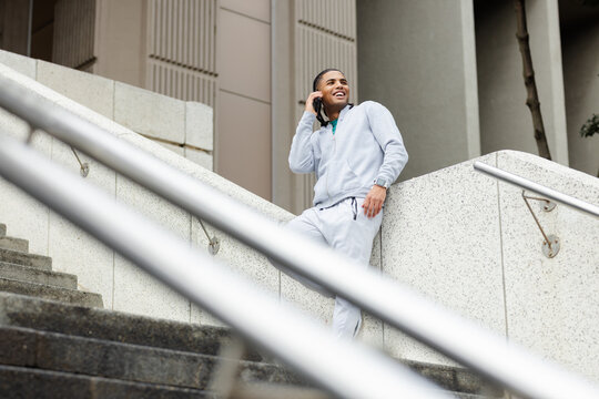 African american man in hoodie leaning by staircase at door holding smartphone smiling