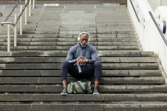 African american man sitting on stairs with headphones, holding bottle checking smartwatch