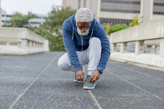 African american man kneeling on asphalt walkway tying shoelaces in blue hoodie and smartwatch