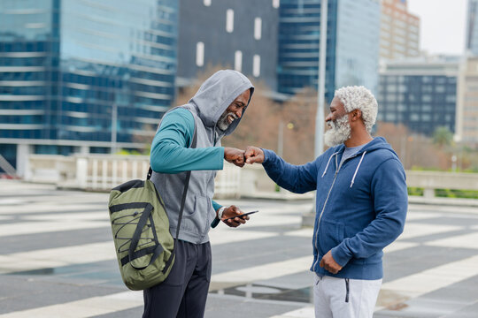 African american male duo fist-bumping on city plaza in sportswear with smartphone and backpack