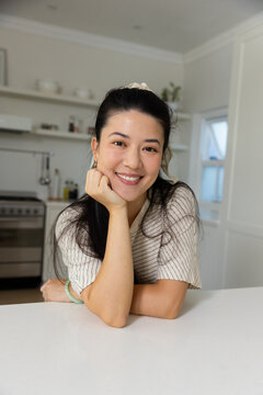 Asian woman leaning on white island countertop near stainless range, smiling, showing bangle