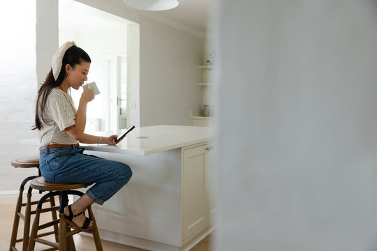 Woman sitting on wooden stool at kitchen island holding ceramic mug checking phone, copy space