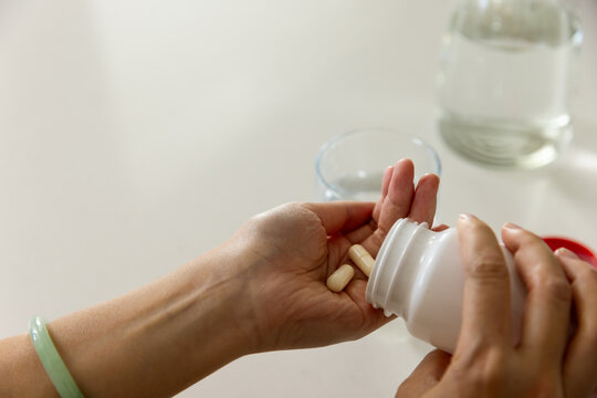 Woman pouring three beige capsules from red-capped bottle into palm on tabletop, jade bangle