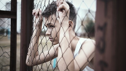 cinematic refugee sad boy stands alone head bowed near the fence, frustrated boy dropped eyes, experience life, have fun and enjoy © vla
