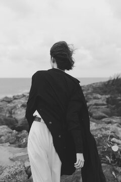 Woman walking along a rocky coastline in a long black coat and white pants, wind swept hair, moody monochrome seascape and distant horizon