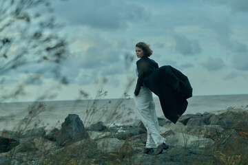 Obraz premium windy coastline scene with a woman walking along rocky shore, flowing coat catching the breeze, ocean horizon in the distance, moody blue tones and natural outdoor atmosphere