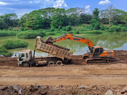 Construction work is taking place by the river; trucks are lifting their trailers to unload soil, and an orange backhoe is parked nearby, assisting in clearing the earth.