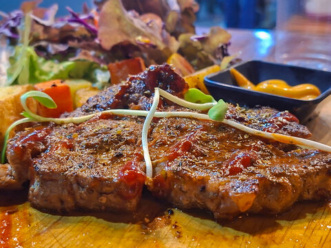 A steak arranged on a wooden cutting board, served with leafy green salad, toasted garlic bread, french fries, and an appetizing yellow sauce in a black cup, resembling cheese or mustard.