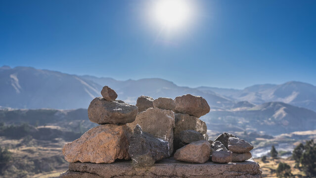 Pyramids of stones are stacked on a boulder. The background is a mountain panorama. The sun is shining in a clear blue sky. Peru. Colca Canyon.