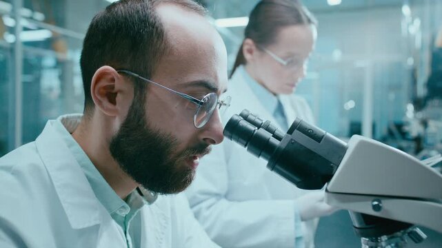 Male scientist observing specimen through microscope and talking to female assistant documenting findings during laboratory research project