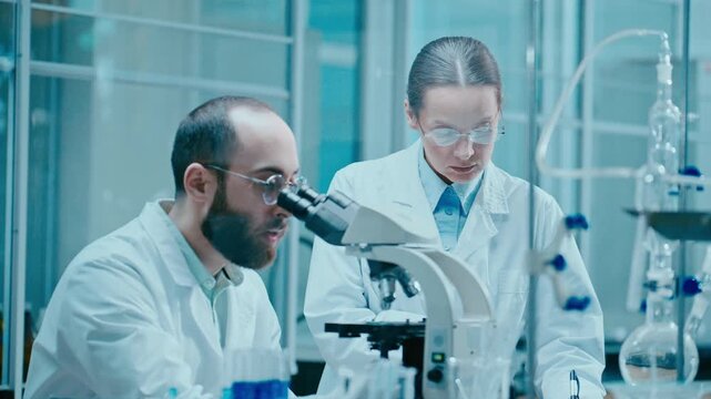 Male scientist observing specimen through microscope and sharing his findings with female colleague writing notes, collaborating on research project in laboratory
