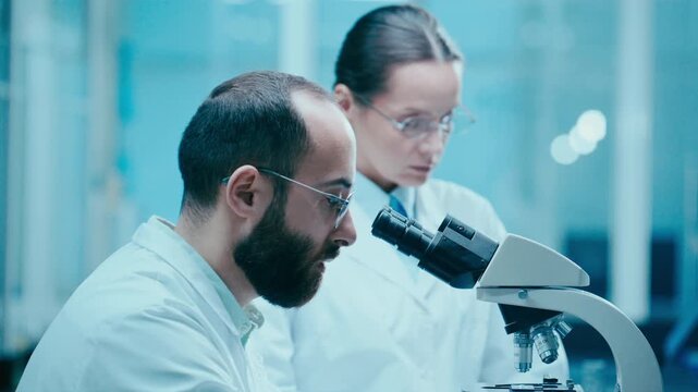 Male scientist using microscope and explaining research results to female colleague as she documenting information in modern laboratory. Rack focus
