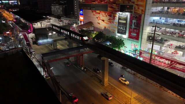 Static aerial night view Bangkok city street with traffic and skytrain passing a shopping mall, illuminated urban buildings, and busy roads, Thailand