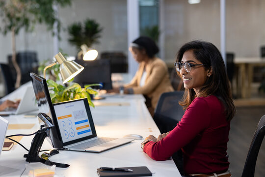 Diverse coworkers working at shared white desk in modern office using laptop and desk lamp