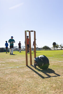 Wooden cricket stumps are standing in dry wicket area and dark green helmet is resting on side