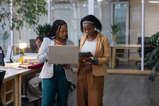 Diverse coworkers collaborating using laptop and tablet in open-plan office with glass partitions