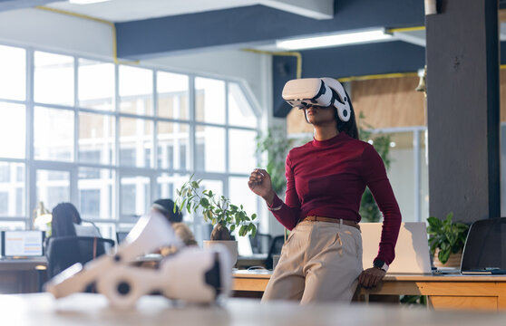 Asian woman wearing white VR headset leaning on wooden desk in open plan office, copy space