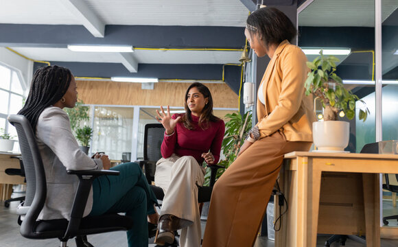 Diverse female coworkers discussing project ideas at open-plan office corner with coffee mug