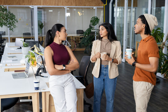 Diverse coworkers leaning on shared desk discussing work plans in office with laptops, mugs