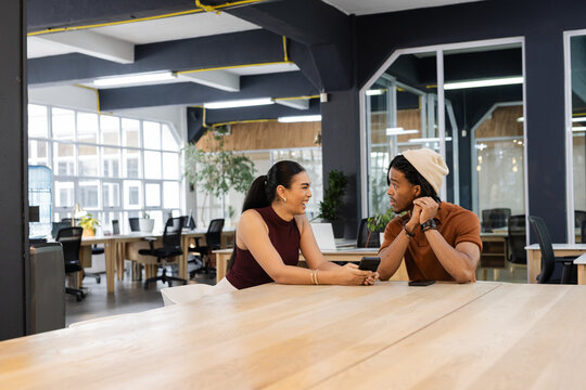 Diverse coworkers leaning over long table speaking on smartphone and adjusting beanie in office