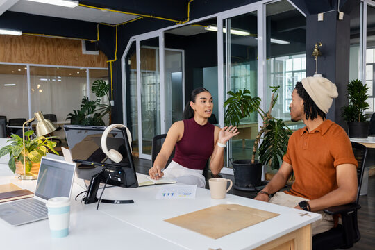 Diverse coworkers gesturing and discussing charts at office desk with open laptop and notebook