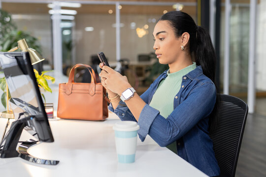 Adult Asian woman checking smartphone sitting at white desk with monitor lamp coffee, copy space