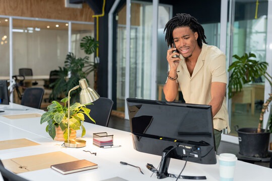 African American man using smartphone leaning toward monitor at office desk with lamp, copy space