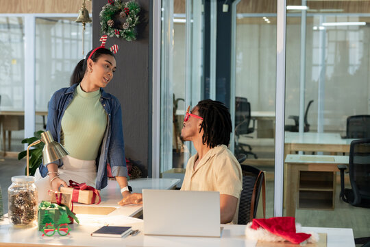 Diverse coworkers exchanging wrapped gift box at desk in office with laptop and reindeer antlers