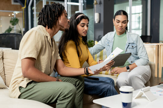 Diverse coworkers discussing printed charts on white table in coworking lounge with tablet