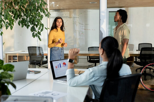 Diverse coworkers collaborating around glass whiteboard in meeting room using laptop and marker