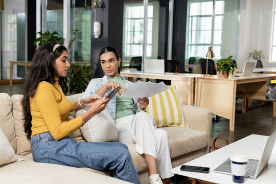 Diverse coworkers sitting on beige sofa in office lounge reviewing tablet and printed documents