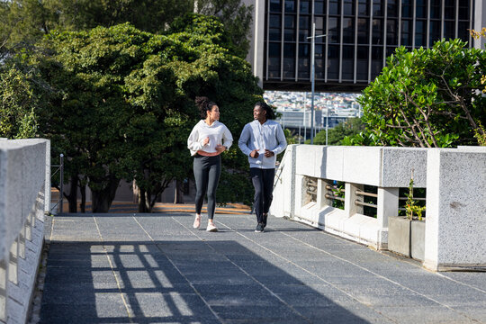 Running in sync on city pedestrian bridge, diverse couple wearing light hoodies, leggings