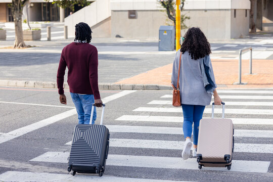 Diverse couple walking across striped crosswalk at street corner pulling suitcases