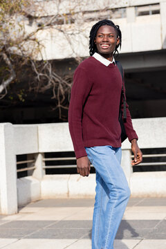 African american man on walkway beside concrete railing in sunlight carrying black backpack