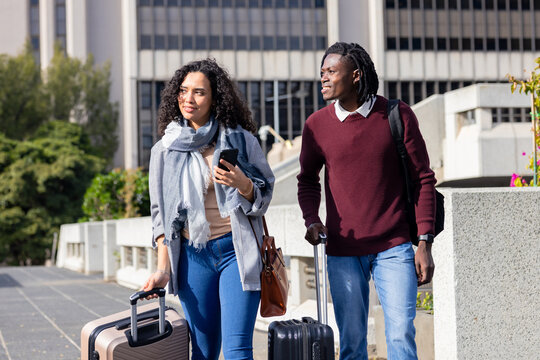 Diverse travelers walking along office walkway pulling rolling suitcases, checking smartphone