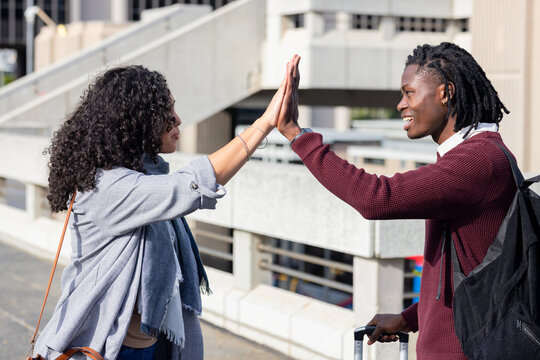 Diverse friends high-fiving on walkway beside parking garage with rolling suitcase, backpack