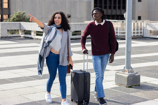 Diverse friends walking at urban plaza pulling suitcase and carrying backpack, crossbody purse