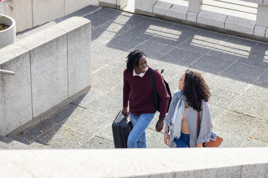 Diverse couple walking up stone terrace, carrying wheeled suitcase, backpack, tan shoulder bag