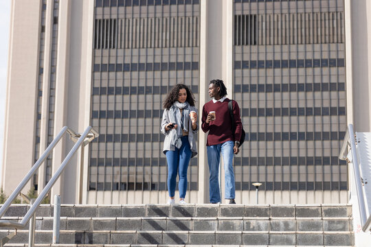 Diverse students walking down campus stairs holding coffee cups, backpacks with smartphone