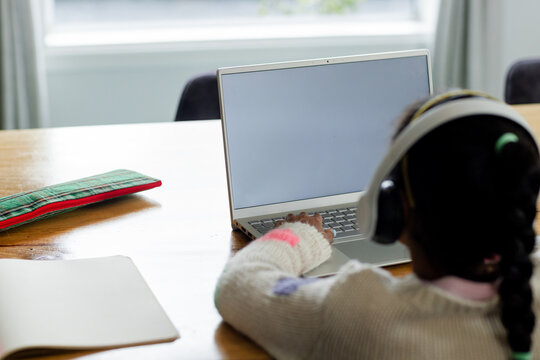 Child typing on laptop at home dining table near window, with notebook and patterned pencil case