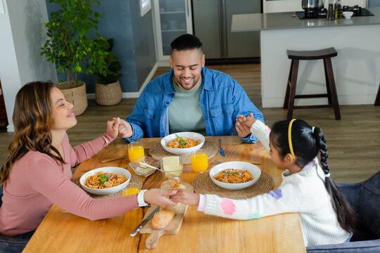 Diverse family praying around wooden dining table with pasta bowls and orange juice in kitchen