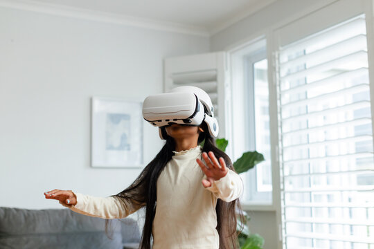 Girl wearing VR headset reaching forward in well-lit living room with gray sofa and white shutters