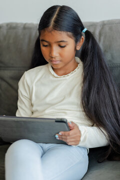 School-aged Asian girl sitting on couch at home holding tablet and focusing on screen