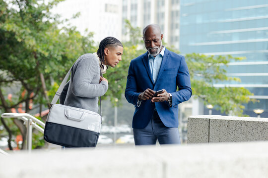 Diverse male coworkers leaning near concrete ledge in city plaza, viewing smartphone screen