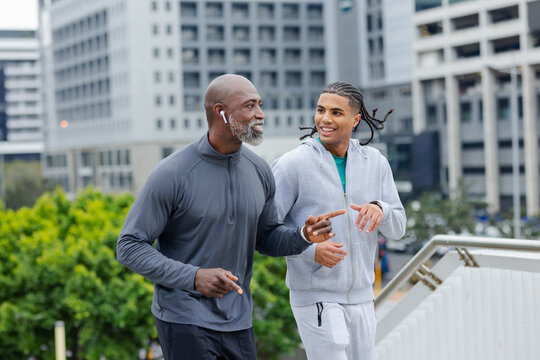 Diverse male friends jogging on elevated city walkway along metal railing using wireless earbuds