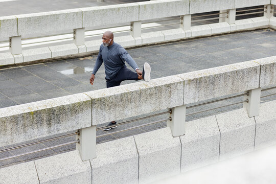 Senior african american man stretching hamstring on urban bridge barrier using earbuds