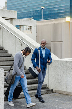 Diverse male coworkers walking down stairs outside office stairs pulling suitcase and messenger bag