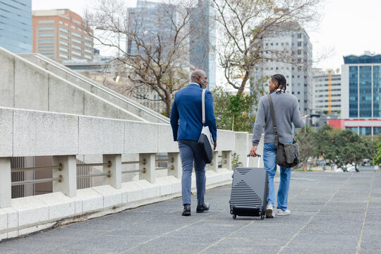 Diverse male coworkers walking beside railing on office walkway holding suitcase and messenger bags